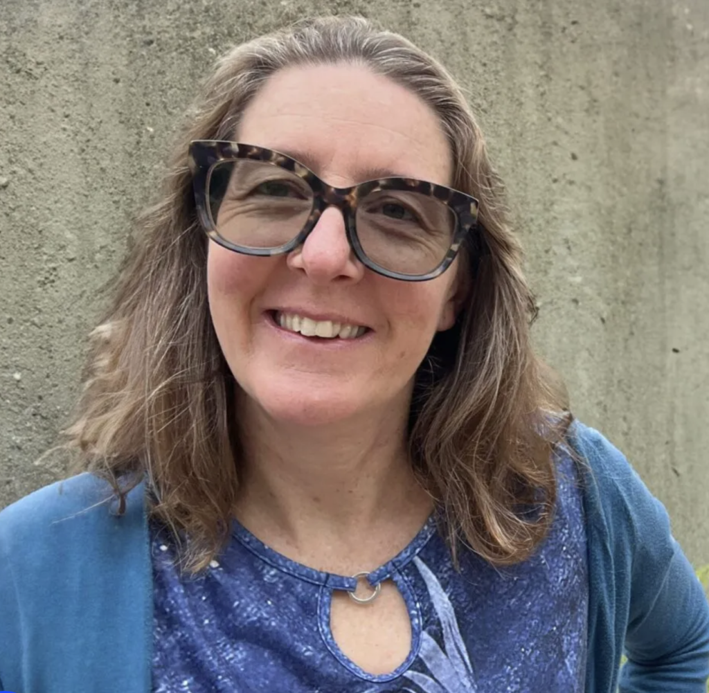 Smiling woman with long wavy hair and large patterned glasses, wearing a blue top and cardigan, standing in front of a concrete wall.